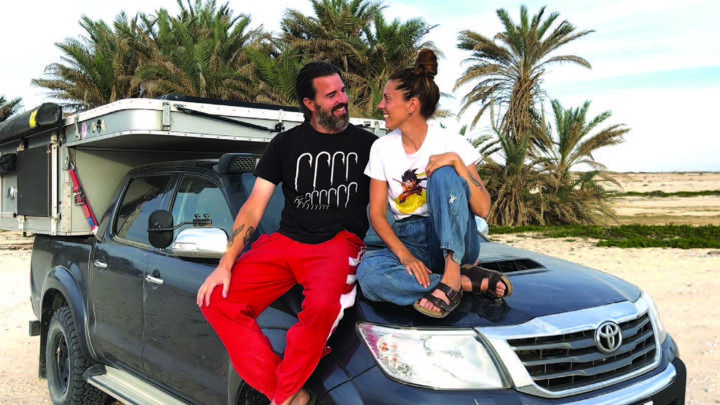 Neus et David assis sur le capot de leur véhicule aménagé, palmiers en arrière-plan — illustration de leur aventure en van life et quête de sens