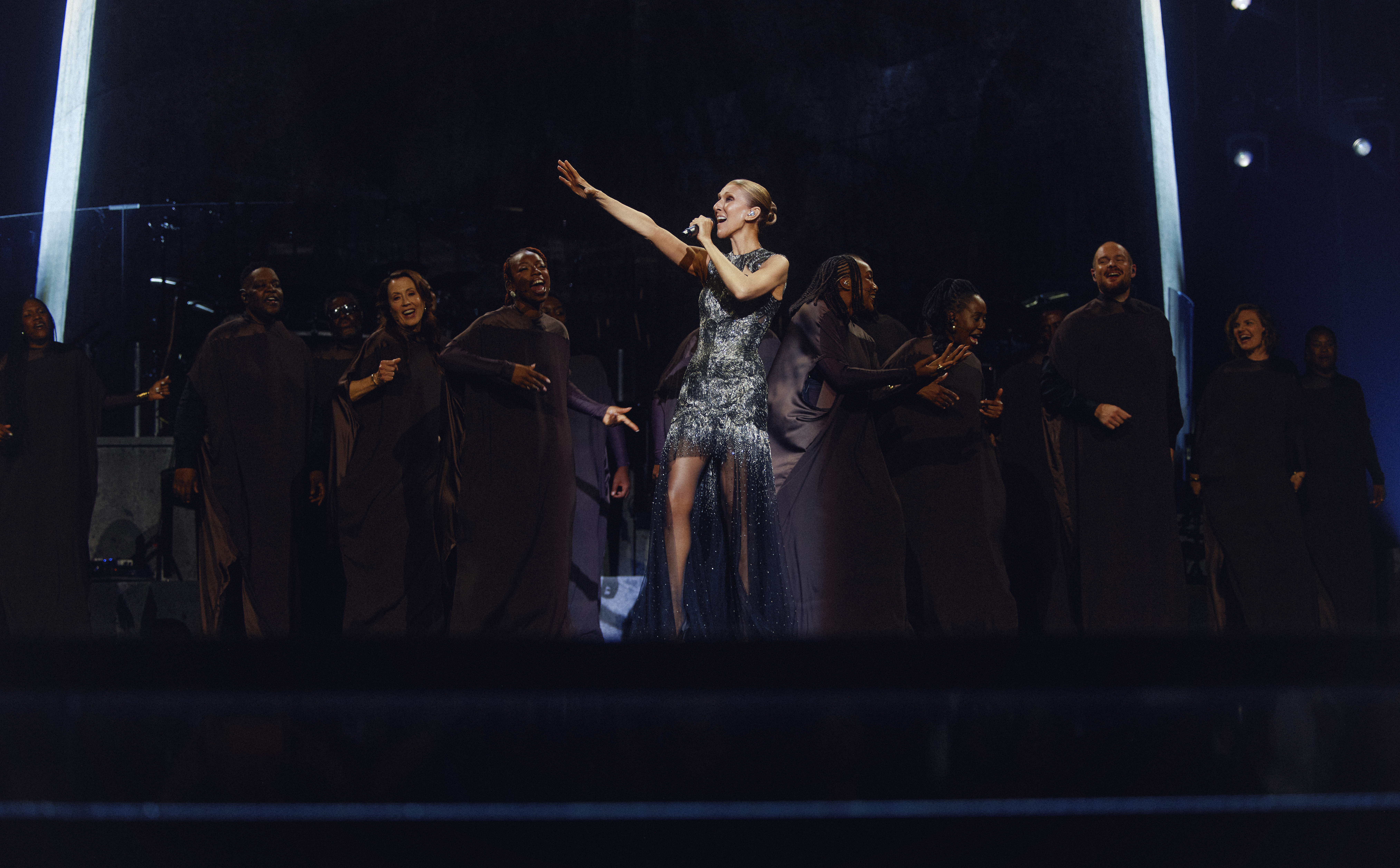 Céline Dion sur scène, entourée de ses choristes, un bras tendu vers le public.