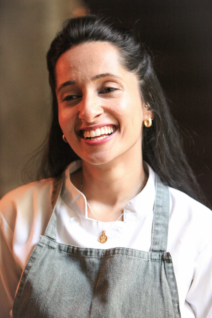Arpitha Savale en veste de cuisine et tablier brun, tenant une poche à douille et une assiette avec un dessert.