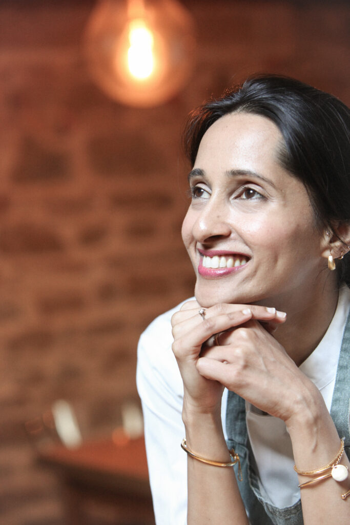 Arpitha Savale souriante, les mains jointes sous le menton, devant un mur de briques.