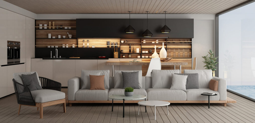 Salon avec canapé gris et fauteuil sombre devant une cuisine ouverte, sous un plafond en bois.