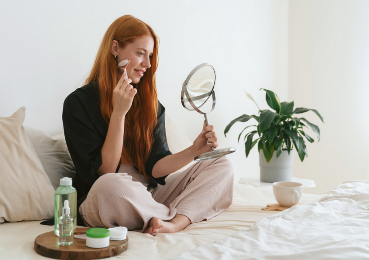 Femme assise sur un lit utilisant un rouleau de massage sur son visage devant un miroir à main.