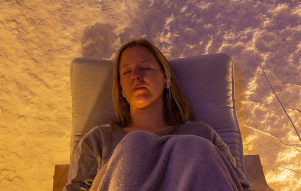 Femme aux yeux fermés, allongée sous une couverture dans un fauteuil, devant une paroi de sel éclairée d’une lumière chaude.